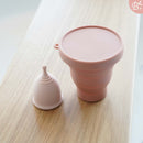 Ein rosa Menstruationsbecher und ein passender faltbarer Sterilisationsbecher auf einer hellen Holzfläche.