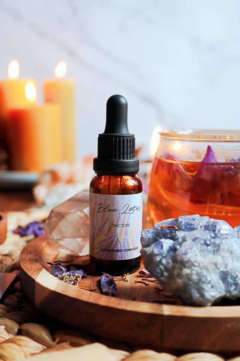 Nahaufnahme einer kleinen Glas-Tropfflasche mit der Aufschrift „Blue Lotus Tincture “ auf einem Holztablett mit einem rauen blauen Kristall, klarem Quarz, losen getrockneten Blütenblättern und einem Glas mit bernsteinfarbener Flüssigkeit, im weichen Hintergrund Kerzen.