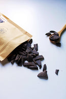 Ein teilweise geöffneter Kraftpapierbeutel mit der Aufschrift „Ayamanu Naturals“ entleert dunkle, unregelmäßig geformte Schokoladenstücke auf eine saubere weiße Oberfläche. Ein Holzlöffel, ebenfalls mit einigen Schokoladenstücken gefüllt, liegt daneben und unterstreicht die natürliche und gesunde Qualität des Produkts.