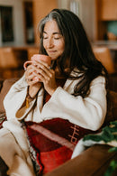 Eine Frau mit graumeliertem Haar und geschlossenen Augen, die auf einem braunen Sofa entspannt und eine rosa Tasse an ihr Gesicht hält.