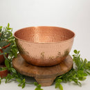 Große, runde gehämmerte Kupferschale, präsentiert auf einem Holzsockel, umgeben von grünen Pflanzen.