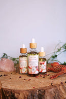 Drei Flaschen "Vanilla" tantrisches Massageöl auf einem Baumstumpf. Die Flaschen, die getrocknete Blüten und Pflanzen enthalten, sind unterschiedlich groß. Ein großer Rosenquarz-Kristall liegt links daneben.