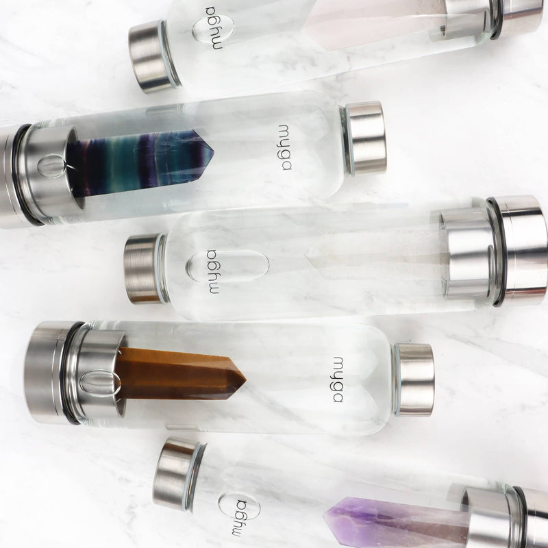  Fünf liegende "myga" Wasserflaschen, jede mit einem einzigartigen Kristalleinsatz: Rosa Quarz, Fluorit, Clear Quarz, Tigerauge und Amethyst, angeordnet auf weißem Marmor.