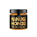 Ein kleines Glas ANUK Manuka Honig MGO 400 mit schwarzem und goldenem Etikett und schwarzem Deckel auf weißem Hintergrund.
