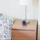 Eine kleine, tiefviolette Amethyst-Geode steht auf einem hellen Nachttisch aus Holz, neben einem Bett mit blau-weiß gemusterter Bettdecke. Eine silberne Tischlampe mit plissiertem weißen Schirm steht ebenfalls auf dem Tisch.