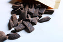  Eine Kraftpapier-Verpackung ist geöffnet und verschüttet dunkle, unregelmäßig geformte Schokoladenstücke auf eine saubere weiße Oberfläche. Etwas Schokoladenstaub ist sichtbar, was auf ein frisches Ausgießen hindeutet.