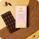 Eine "delikao Feel Good" 70% Zartbitterschokolade mit Haselnusspraline und ihre Verpackung, platziert auf einem diagonalen Band aus kakaofarbener Erde oder Sand, auf einem zweifarbigen beigen und gelben Hintergrund.