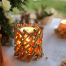 Ein rustikaler Kerzenhalter aus Holz, dessen warmes Licht durch die gekreuzten Stäbchen scheint, steht auf einem weißen Tisch neben einem Blumenarrangement.Ein rustikaler Kerzenhalter aus Holz, dessen warmes Licht durch die gekreuzten Stäbchen scheint, steht auf einem weißen Tisch neben einem Blumenarrangement.