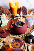 Ein Teetisch mit einer roten Teekanne, einem Glas mit Kräutertee, Kerzen, einem Amethystkristall, einer Teepackung und einer Tarotkarte, die "The High Priestess" zeigt.