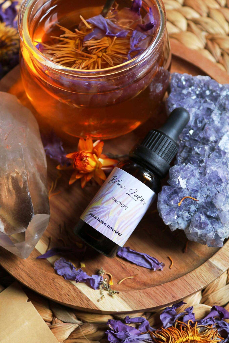 Eine Flasche "Blue Lotus Tinktur" ruht auf blauen Kristallen auf einem Holztablett, neben einem großen klaren Quarzkristall und einem Glas bernsteinfarbener Flüssigkeit mit violetten Blütenblättern. Getrocknete Lotusblütenblätter und andere botanische Elemente sind um das Tablett verstreut, das auf einer gewebten Matte liegt.