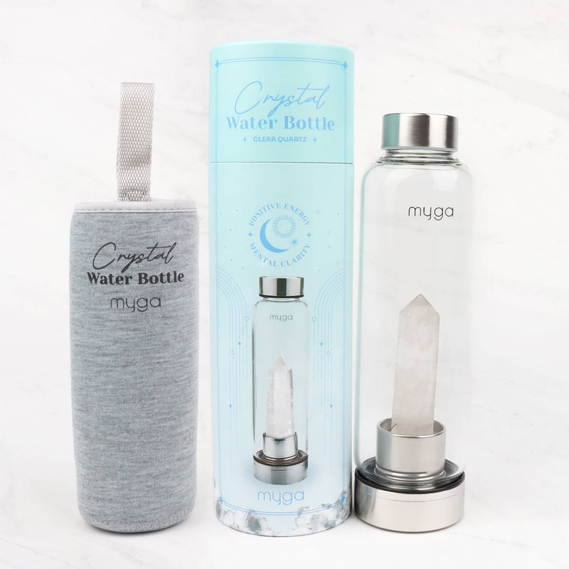 Eine Wasserflasche in einer grauen Stoffhülle, eine hellblaue zylindrische Box mit der Aufschrift "Crystal Water Bottle + Clear Quartz" und eine durchsichtige Glaswasserflasche mit einem Kristall darin, alles auf einer weißen Marmoroberfläche.