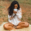 Eine Frau mit langen, dunklen Locken sitzt im Schneidersitz auf einem Fellteppich und trinkt aus einer weißen Tasse.