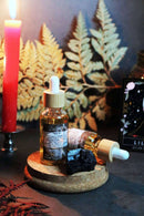 Eine Ritual-Altarszene mit einer brennenden roten Kerze, zwei Pipettenflaschen Ritualöl (eine stehend, eine liegend) mit Bambusverschluss, einem schwarzen Stein und einer "Lilith"-Orakelkarte. Im Hintergrund sind große getrocknete gelbe Farne und eine dunkle, mystische Stimmung zu sehen.