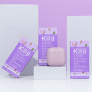 Seifenschachteln mit der Aufschrift „Koji White Kojic Acid & Hyaluronic Skin Brightening Soap“ und ein rosa Seifenstück werden vor einem pastellvioletten Hintergrund präsentiert.