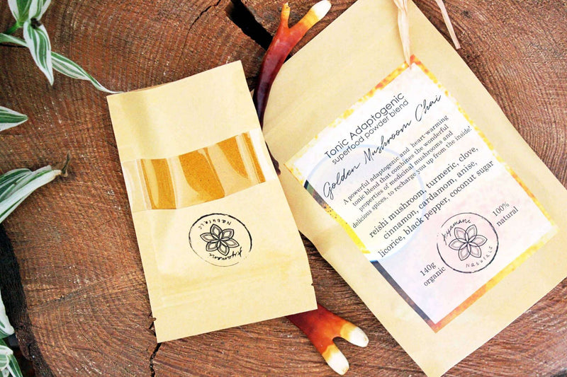 Eine Nahaufnahme von zwei Kraftpapierbeuteln mit "Golden Mushroom Chai", die auf einer dicken Scheibe eines Baumstamms liegen. Die Beutel sind von grün-weiß gestreiften Blättern und getrockneten Reishi-Pilzen umgeben, was eine natürliche, erdige Ästhetik erzeugt.