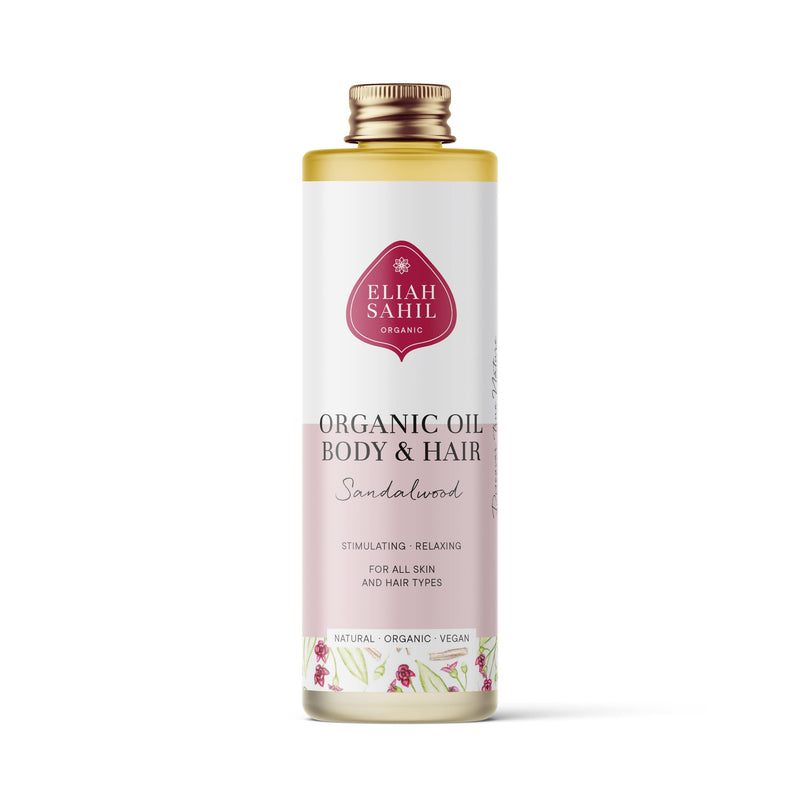 Produktfoto einer hohen weißen Flasche „Eliah Sahil Organic Oil Body & Hair Sandalwood“ mit einem hellrosa Etikett, das das Öl als stimulierend und entspannend für alle Haut- und Haartypen beschreibt.