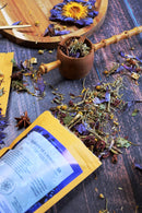 Eine Nahaufnahme von losem Kräutertee, der aus einer geöffneten Tüte und einem hölzernen Löffel auf einer dunklen Holzoberfläche verschüttet wurde, mit getrockneten Blütenblättern und Sternanis daneben.