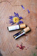 Eine getrocknete blaue Lotusblüte mit gelbem Staubgefäß liegt neben zwei Roll-on-Flaschen mit Chakra-Öl und Holzdeckeln, umgeben von trockenen rosa Blütenblättern, auf einer Holzoberfläche.