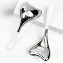 Zwei herzförmige silberne Gesichtsmassagegeräte mit weißen Griffen auf einer weißen Fläche.