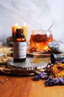 Eine kleine, offene Bernsteinfarbe-Flasche „Blue Lotus Tincture “ steht auf einem runden metallischen Untersetzer neben einer Pipette und getrockneten lila Lotusblüten. Brennende Kerzen und ein Glas mit bernsteinfarbenem Tee sind im weichen Hintergrund sichtbar.