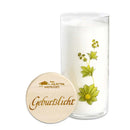 Eine hohe weiße Kerze in einem mit Blumenmuster verzierten Glashalter, neben einem runden Holzdeckel mit der Aufschrift „Geburtslicht“.