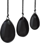 Ein Set aus drei glatten, polierten Yoni-Eiern oder Anhängern in verschiedenen Größen, geschnitzt aus glänzendem schwarzem Obsidian und an verknoteten schwarzen Schnüren hängend.