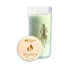 Eine hohe Glaskerze mit grünem Wachs und Blumenillustration, neben einem Holzdeckel mit der Aufschrift „Vergebung“ und „Achtsamkeit“.